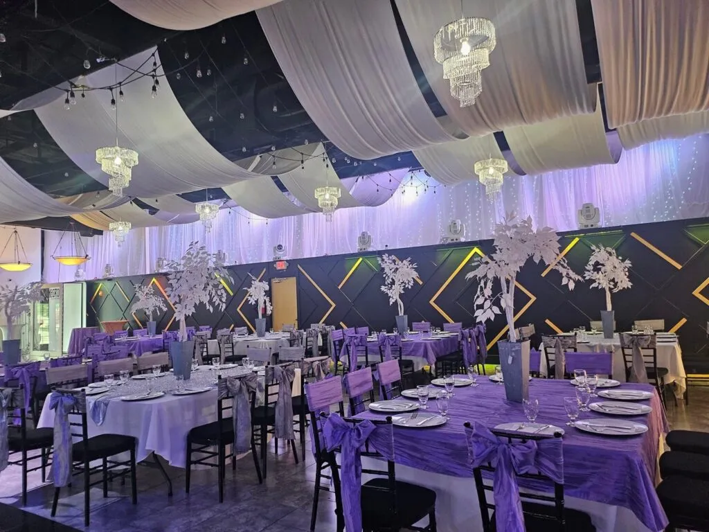 Mesas de banquete en lavanda y blanco bajo un techo con telas y candelabros.